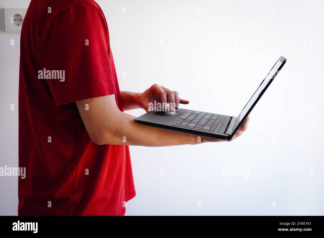 Giovane investitore di sesso maschile in maglietta rossa in piedi in una stanza con pareti bianche e laptop, che naviga su Internet Foto Stock