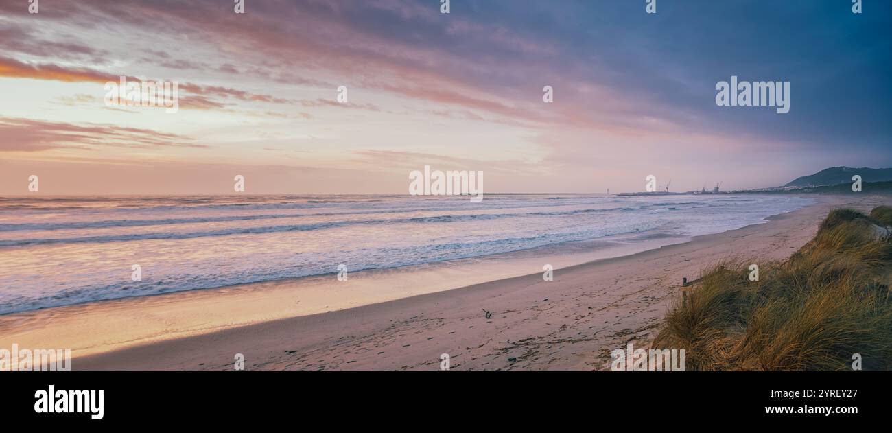 Onde che si infrangono sulla riva della spiaggia di rodanho all'alba con cielo color pastello e gru industriali a viana do castelo, portogallo Foto Stock