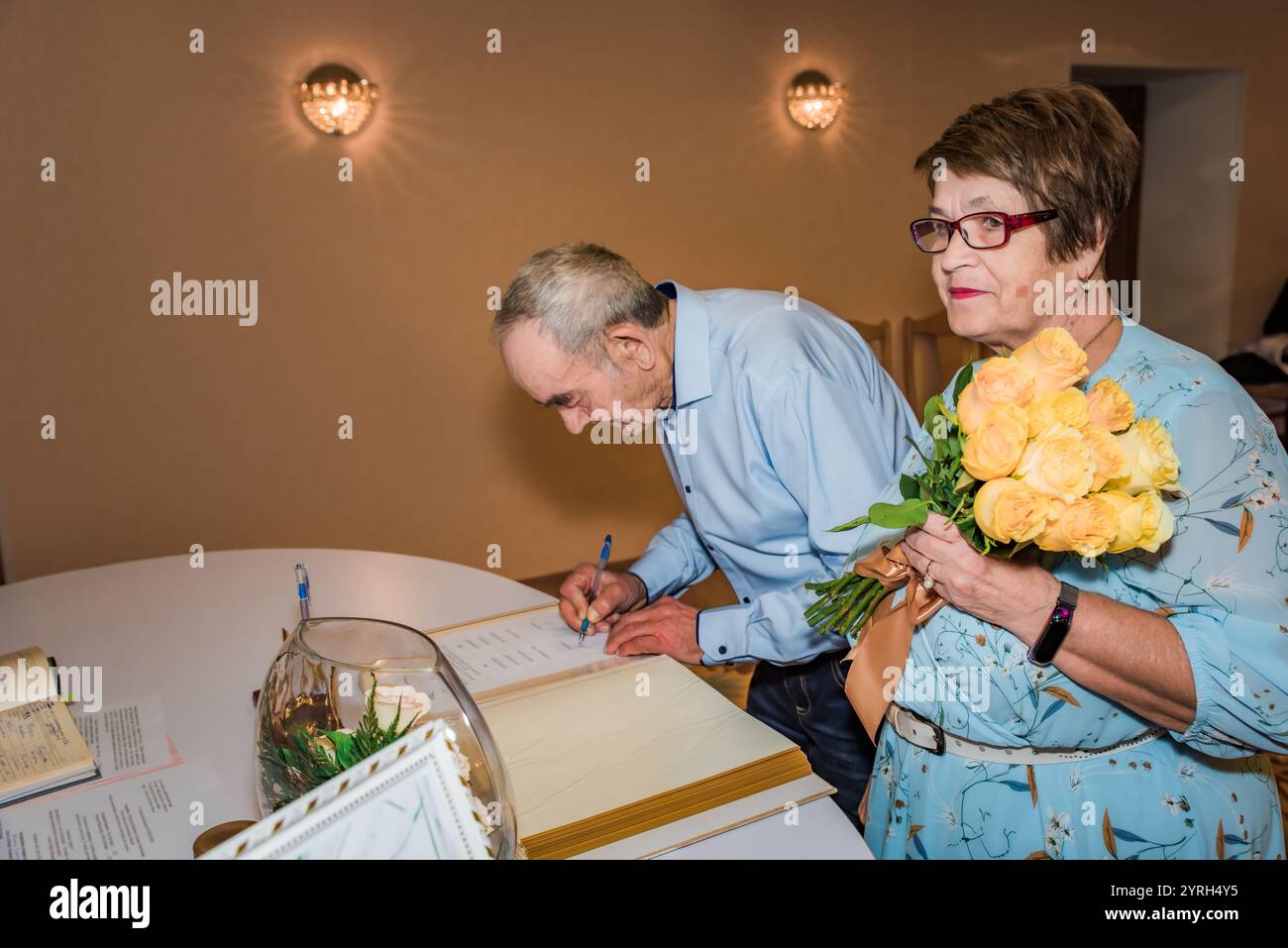 Una coppia di anziani firma documenti in un ufficio di registro in Russia, per celebrare il loro 50 ° anniversario di matrimonio. La donna regge un bouquet di rosa gialla Foto Stock
