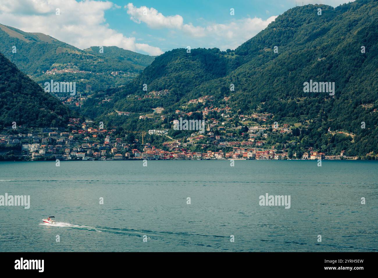 Il motoscafo crea una scia sul lago di como, con un affascinante villaggio incastonato tra montagne lussureggianti sotto un cielo blu con nuvole bianche. Preso da Lezzeno Foto Stock