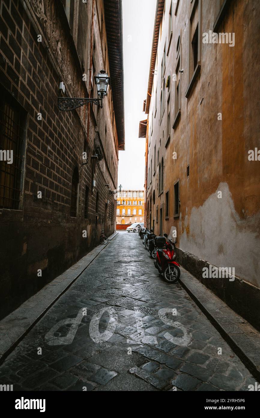 Gli scooter parcheggiati fiancheggiano uno stretto vicolo acciottolato a firenze, in italia, che conduce verso una strada più grande e luminosa e vecchi edifici Foto Stock