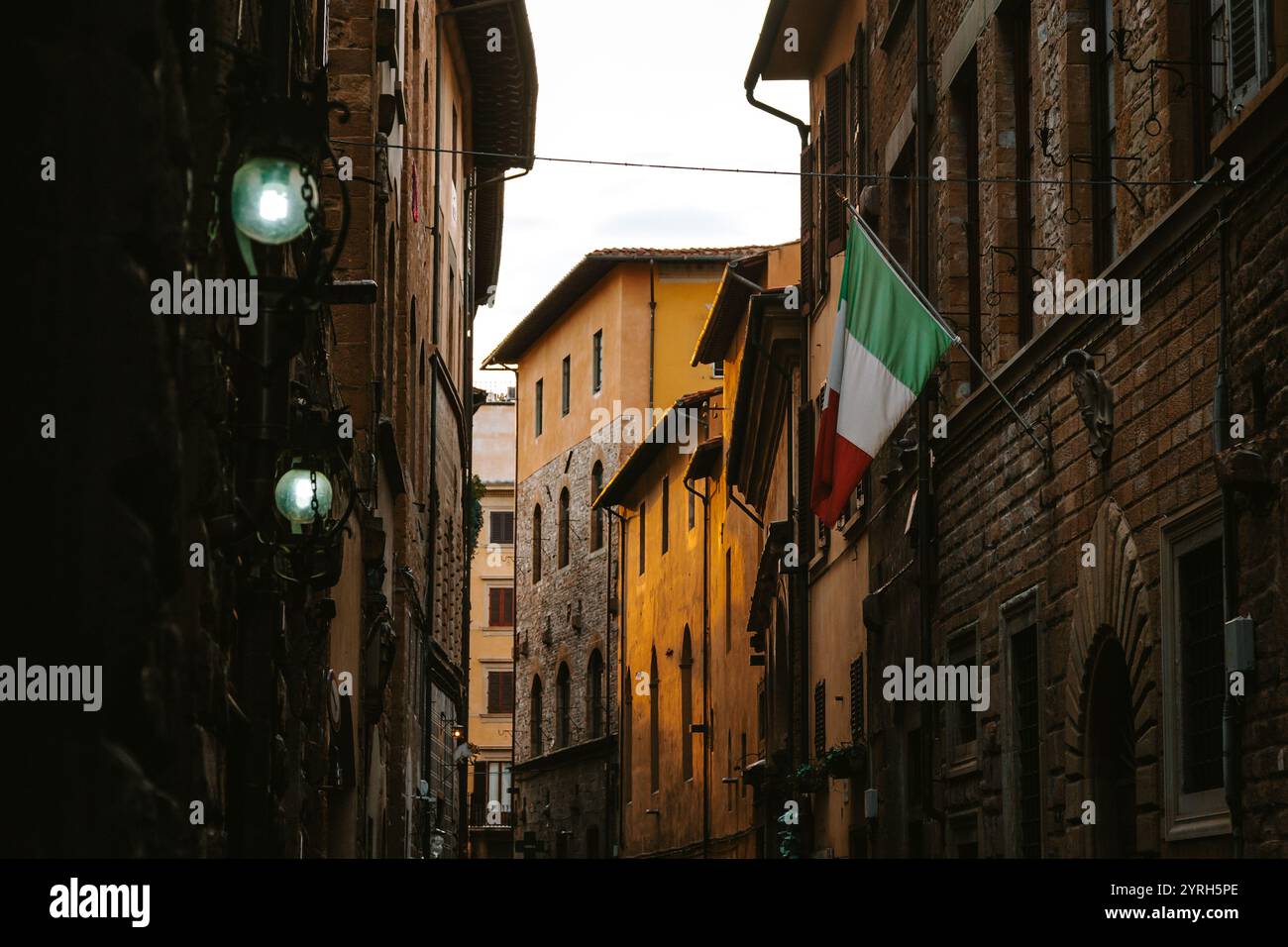 Calda luce del tramonto che illumina edifici colorati in una stretta strada di firenze, con una bandiera italiana appesa a un edificio e lampade d'epoca Foto Stock