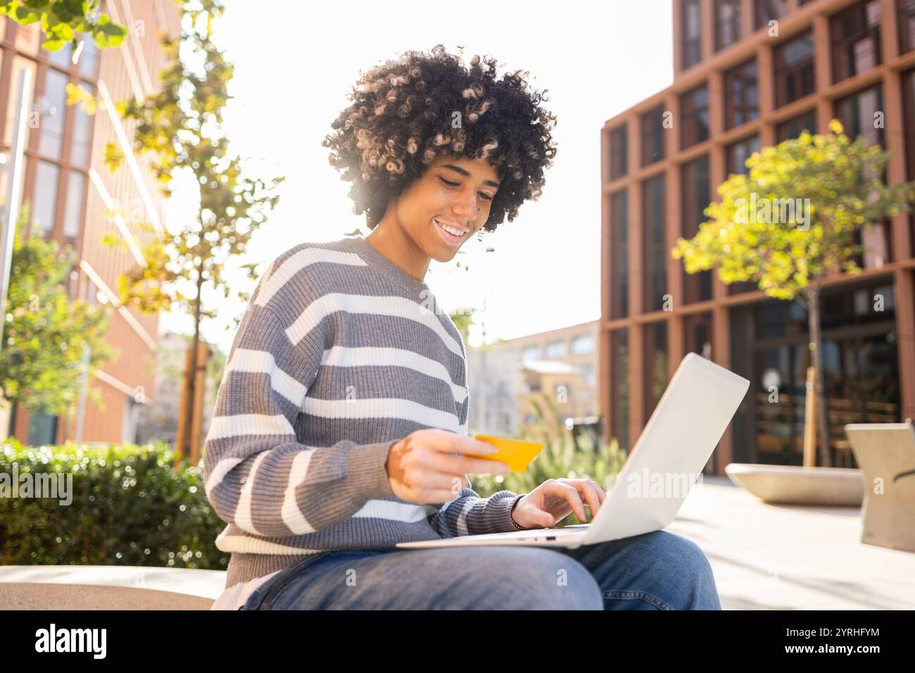 Una donna di razza mista con vitiligine sorride mentre usa il suo laptop all'aperto in un campus universitario, possiede una tessera, che mostra un momento di relax e.. Foto Stock