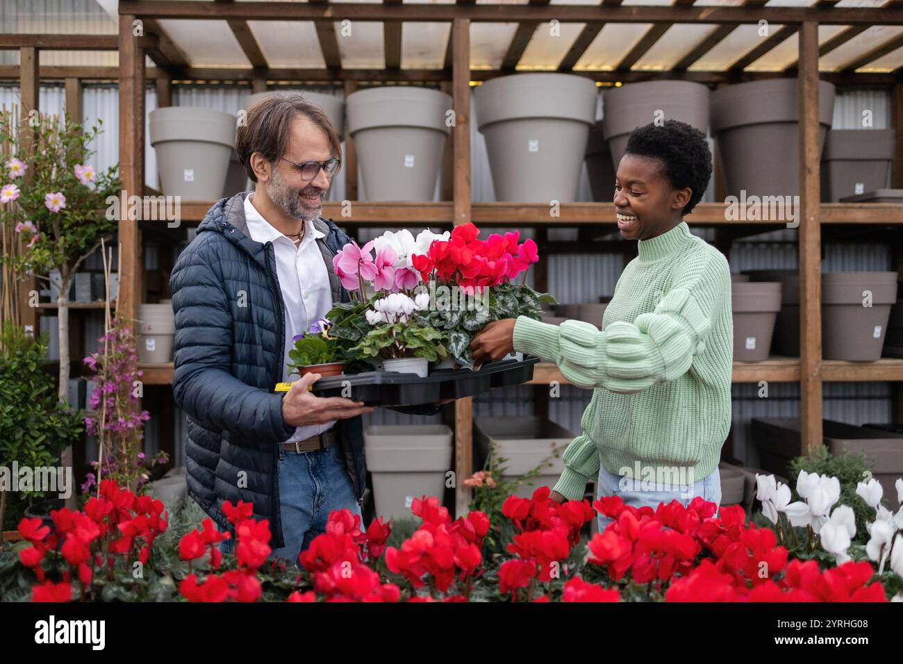 Due clienti sorridono calorosamente mentre tengono in mano un vassoio di fiori colorati in un vivace negozio di giardini. Una serie di vasi e fiori vivaci creano un vivace atmo Foto Stock