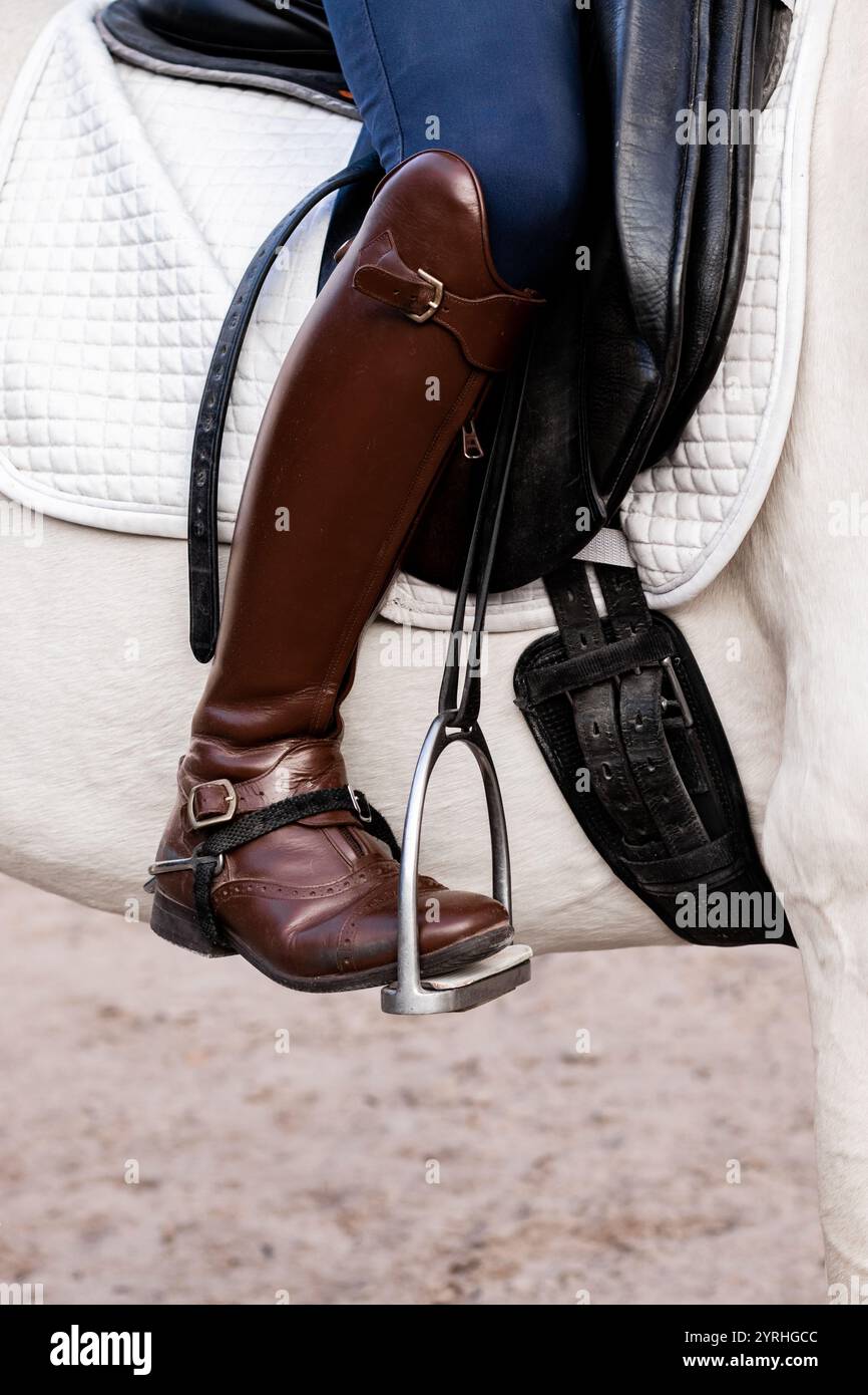 Primo piano di uno stivale marrone in pelle di un pilota in una staffa di metallo su un cavallo bianco, che mette in risalto l'equipaggiamento equestre. La sella bianca e la sella scura Foto Stock