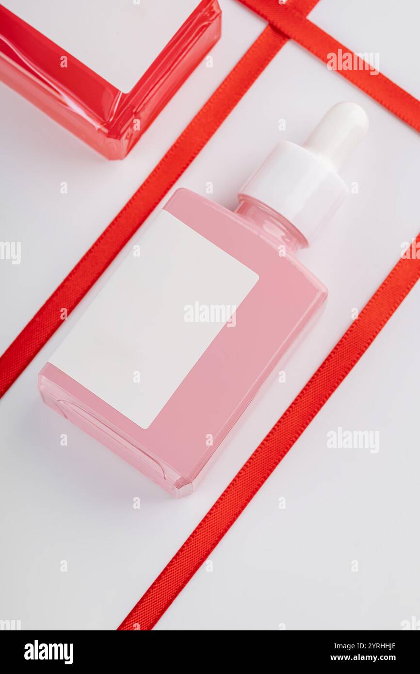 Confezione estetica con contagocce rosa e rosso con etichetta vuota, decorata con nastri rossi vivaci su sfondo bianco, ideale Foto Stock