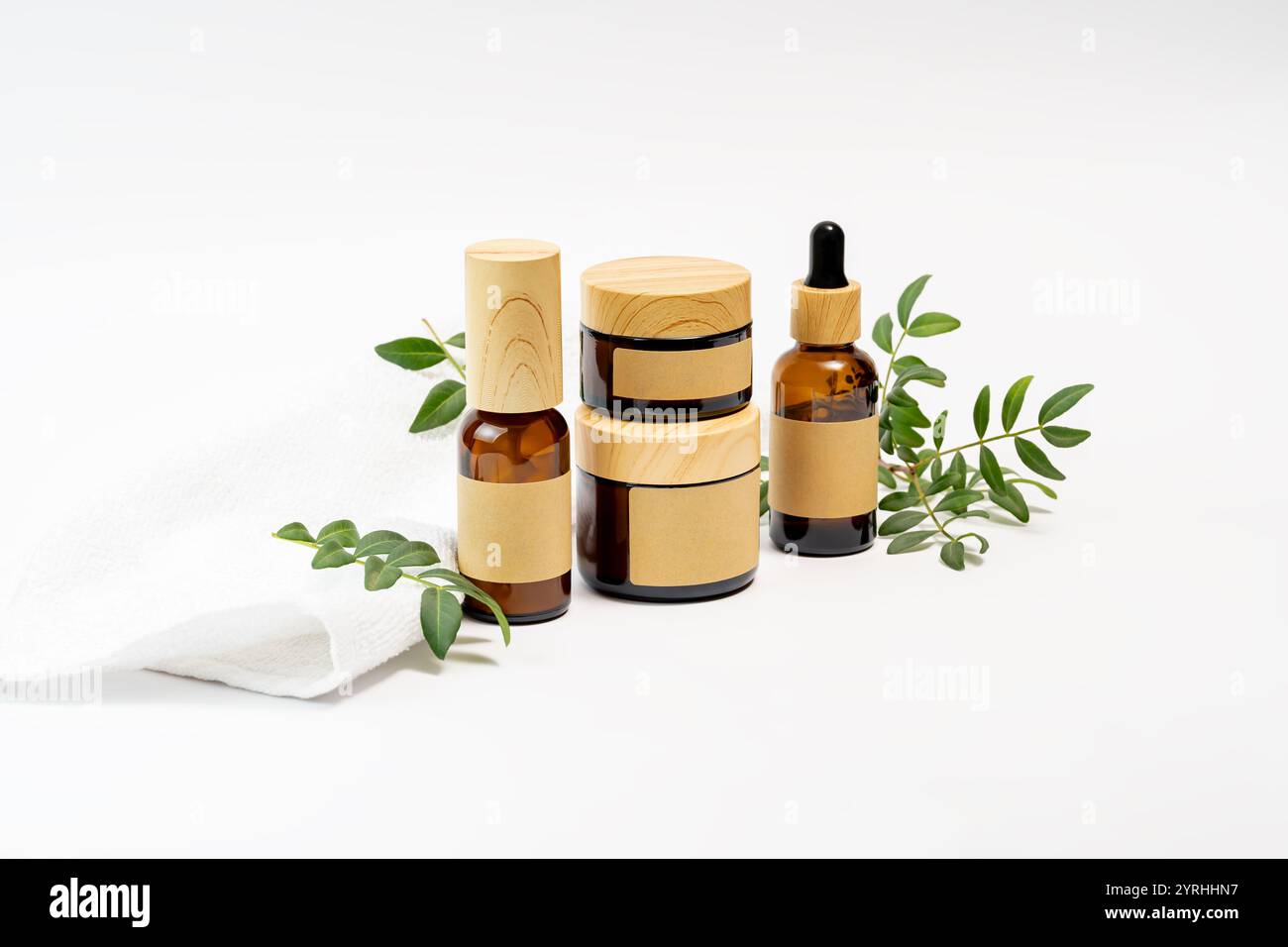Un set di bottiglie per cosmetici eco-compatibili con coperchi ed etichette in legno, disposte con un asciugamano bianco e rami verdi su sfondo bianco, che incarnano un Foto Stock