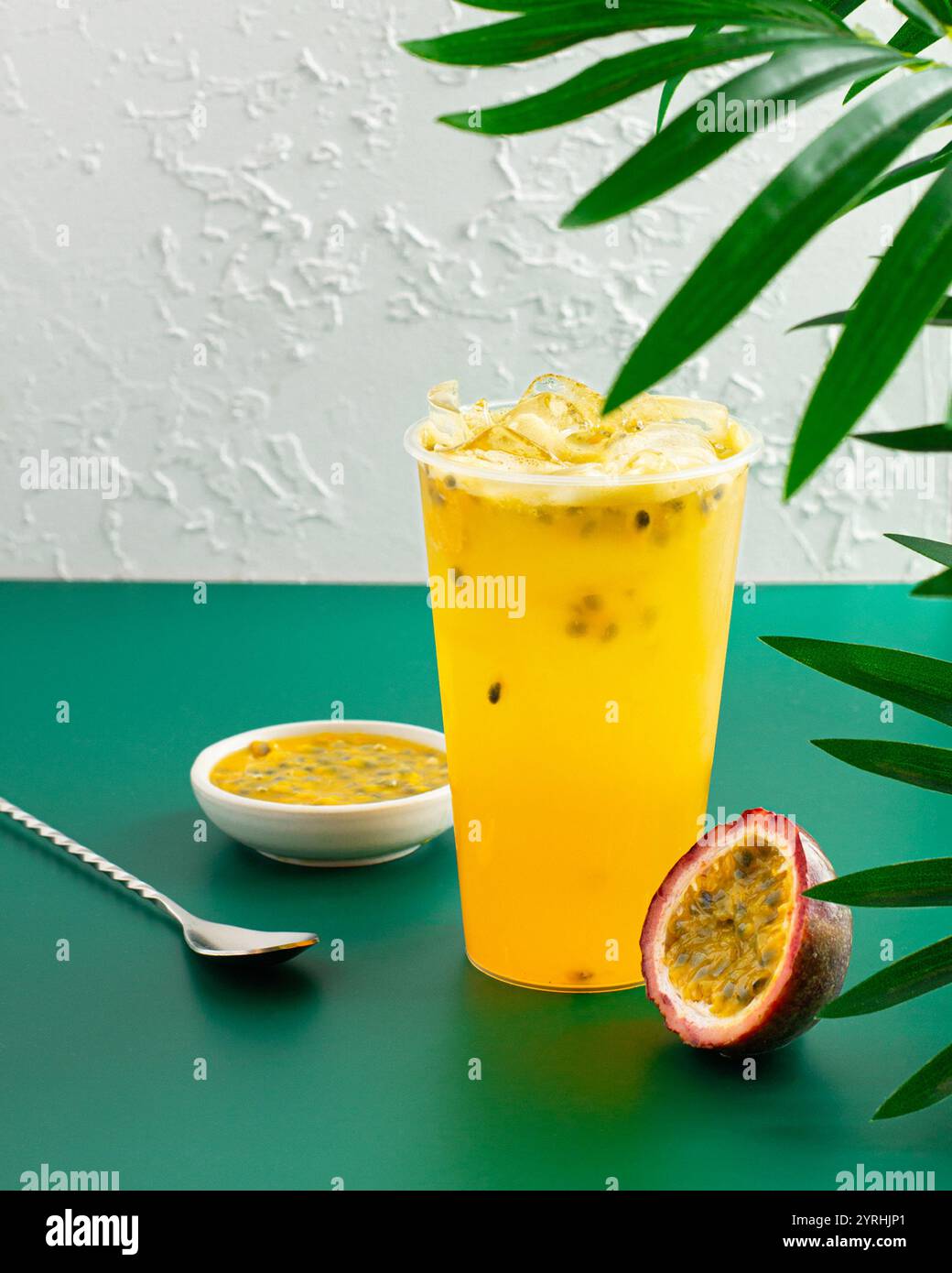 Una vivace tazza di succo di frutta della passione ghiacciato, accompagnata da frutta a fette e una piccola ciotola di polpa, appoggiata su una parete testurizzata e fogliame verde per una t Foto Stock