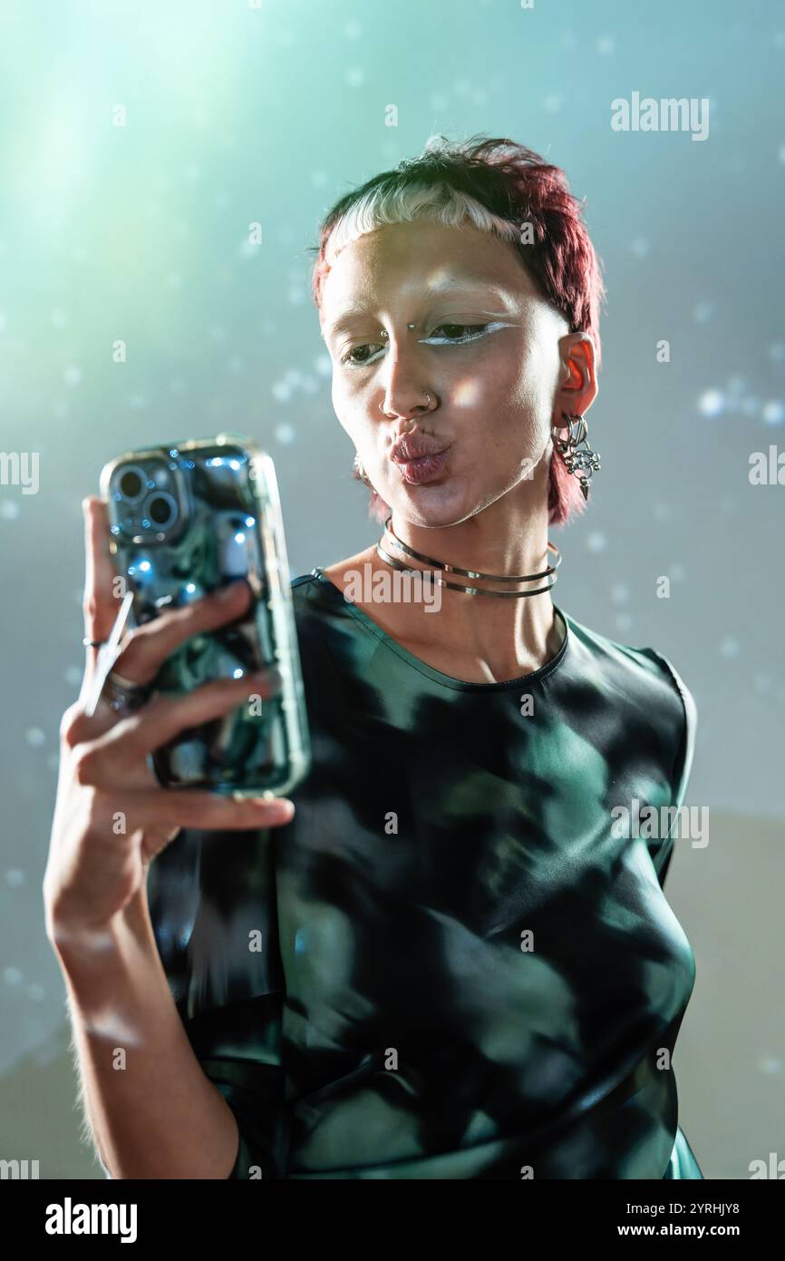 Una giovane donna con un'acconciatura moderna e audace si fa un selfie giocoso, schiacciando le labbra con il suo look futuristico su uno sfondo tenue e luminoso Foto Stock