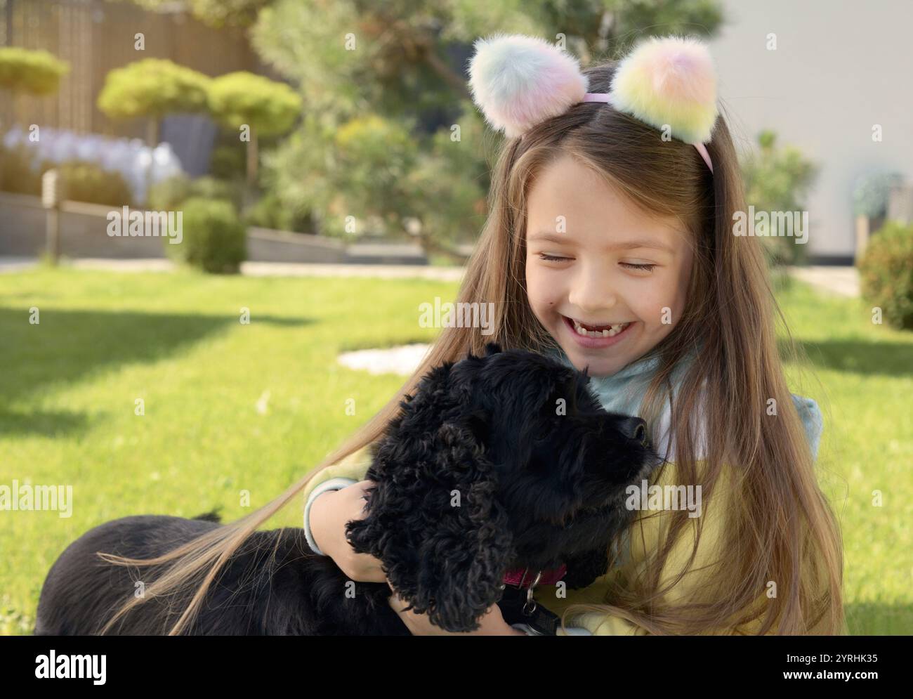 Una giovane ragazza allegra che indossa un soffice archetto color pastello abbraccia felicemente un cane nero in un parco giochi soleggiato il verde brillante li circonda, addi Foto Stock