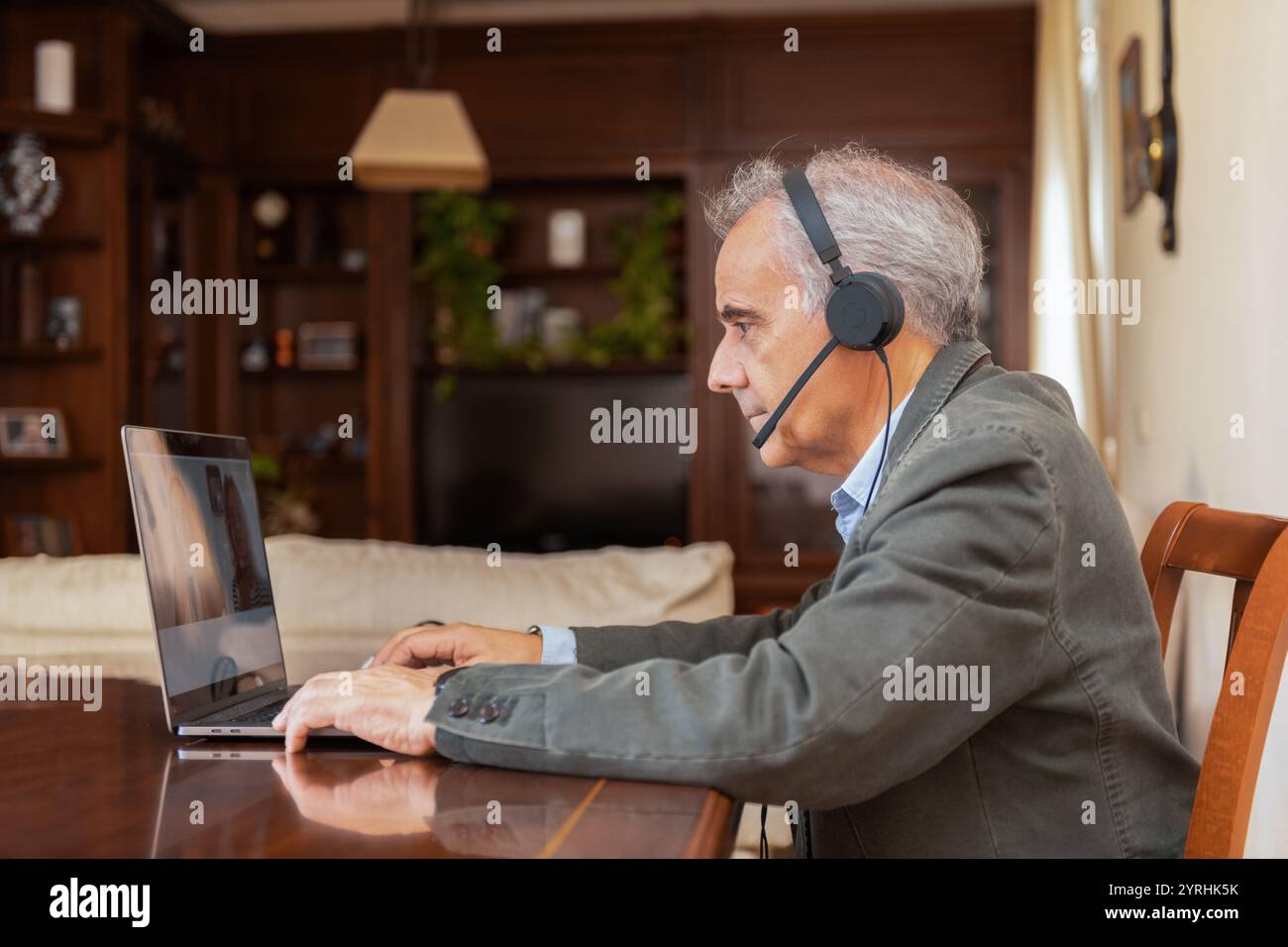 Un uomo maturo con i capelli grigi, indossa una cuffia, effettua una videochiamata su un computer portatile da casa. È concentrato e attento, mostrando una dinam di lavoro a distanza Foto Stock
