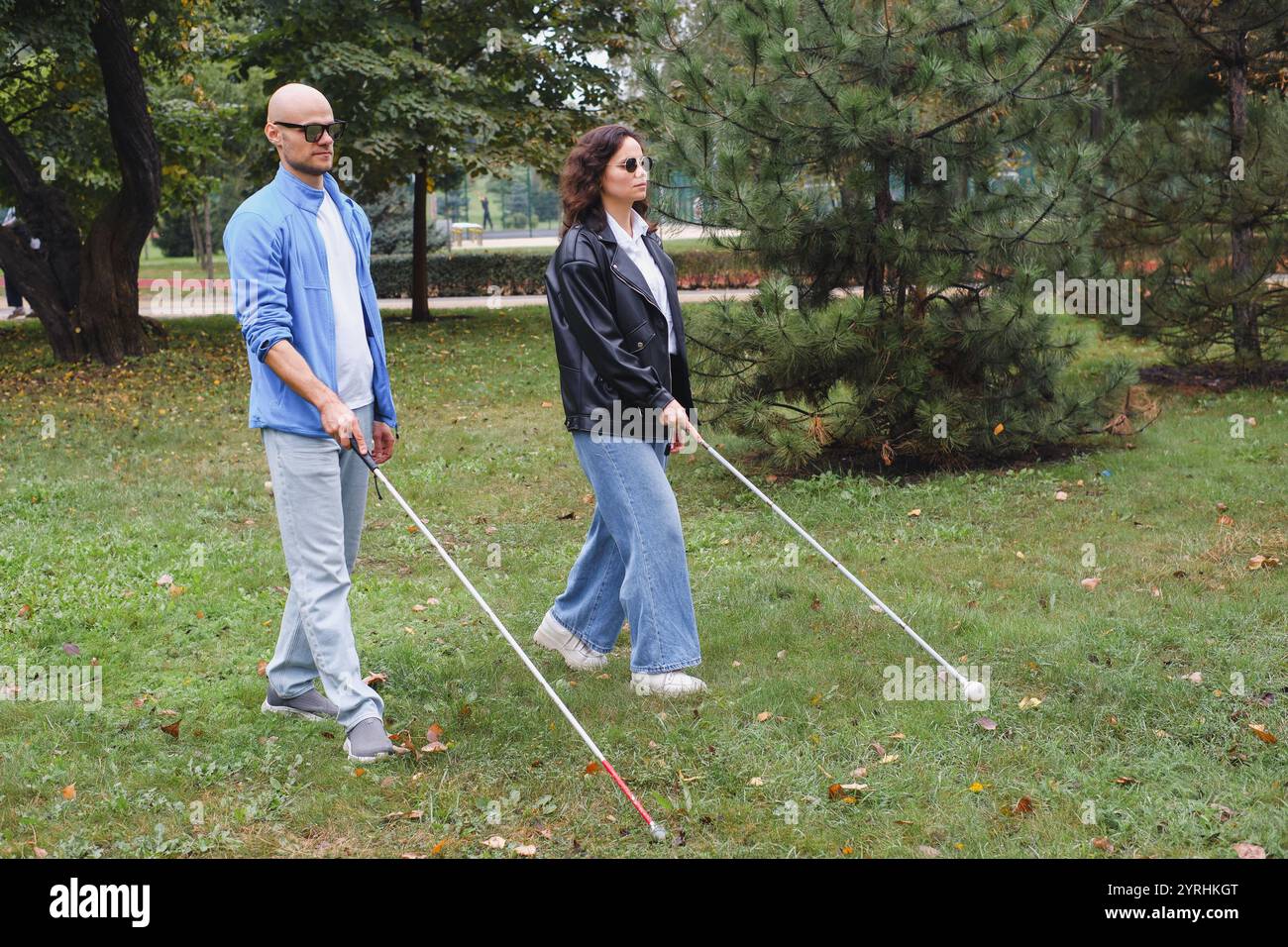 Due persone con disabilità visive camminano attraverso un parco, utilizzando canne bianche per la navigazione. Circondati da lussureggianti alberi verdi, godono di una posizione tranquilla Foto Stock