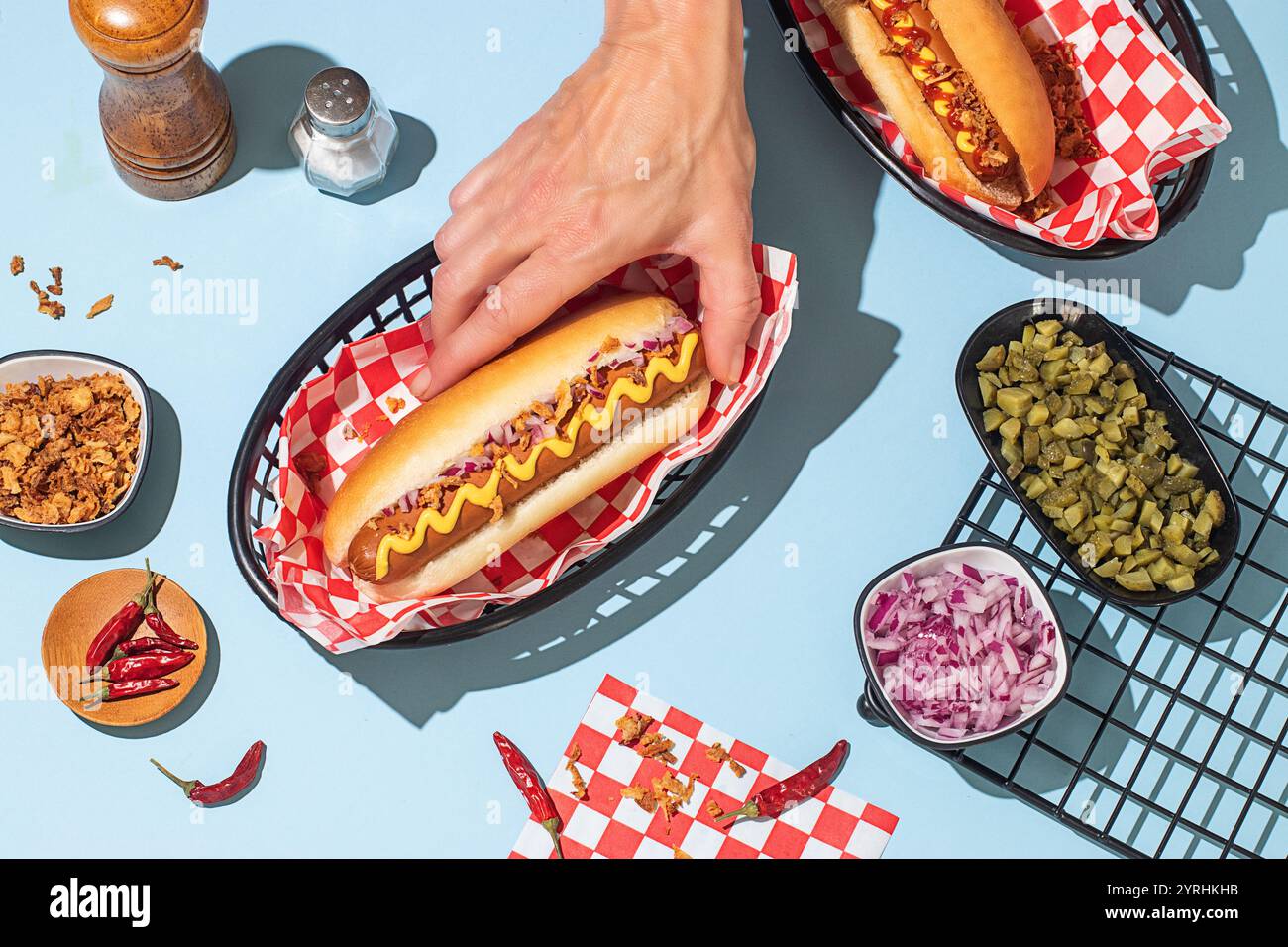 Un primo piano di mani che tengono in mano un hot dog con senape, circondato da condimenti colorati su uno sfondo blu brillante Foto Stock