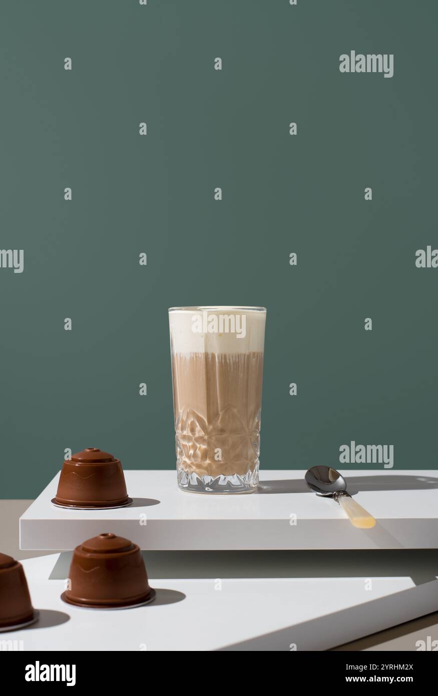 Un alto bicchiere di caffè ricoperto di crema di latte, su un tavolo moderno con capsule di caffè e un cucchiaio nelle vicinanze, perfetto per immagini a tema caffetteria Foto Stock