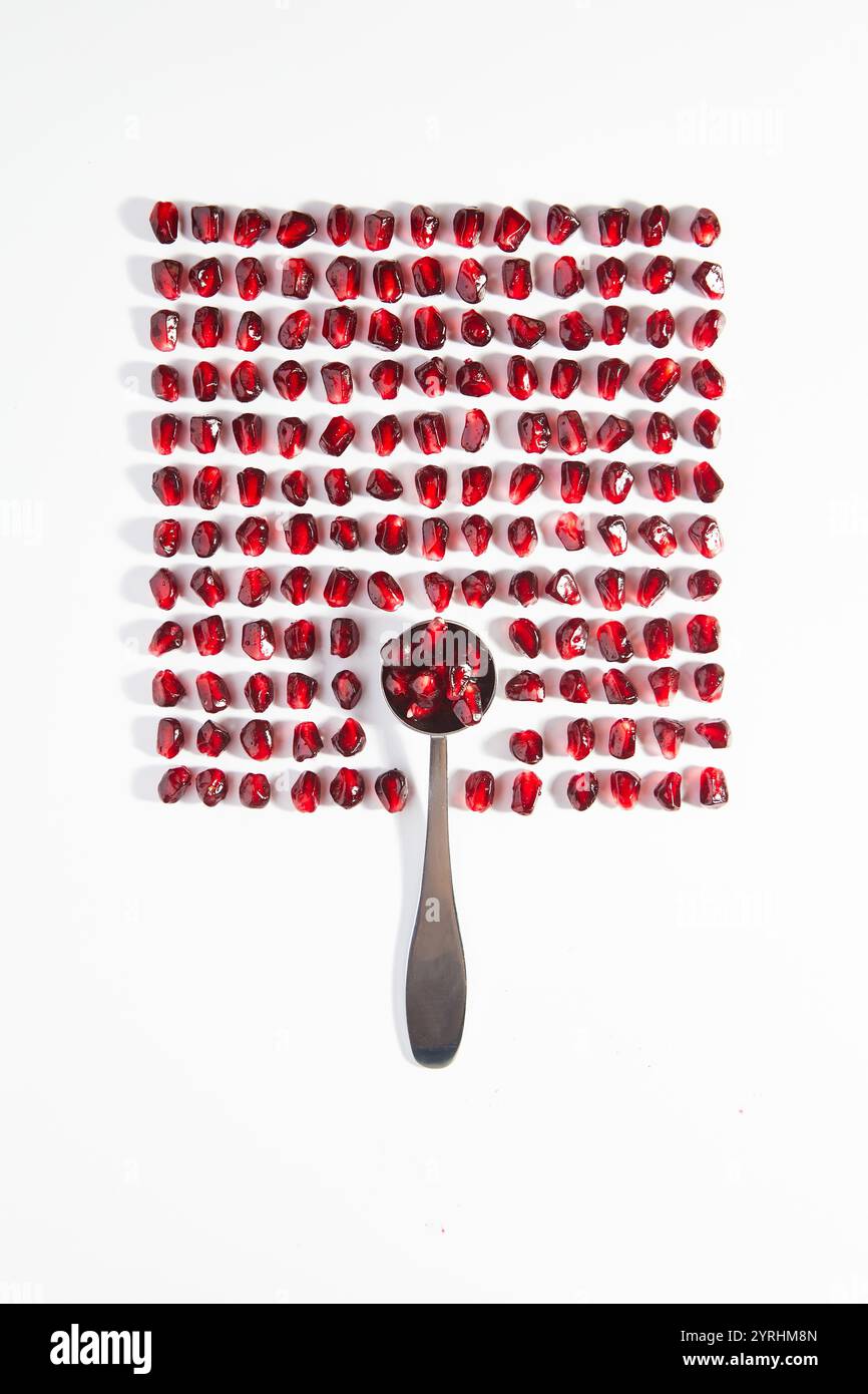 Una disposizione visivamente sorprendente di semi di melograno rosso ordinatamente organizzati in una griglia, con un cucchiaio che tiene alcuni semi, adagiato su un backgrou bianco pulito Foto Stock