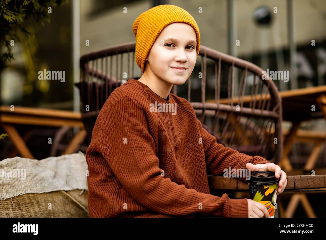 Giovane che indossa un maglione marrone e un cappello in maglia arancione, gustando una bevanda calda all'aperto scena urbana autunnale con colori caldi e accoglienti che catturano la stagione Foto Stock