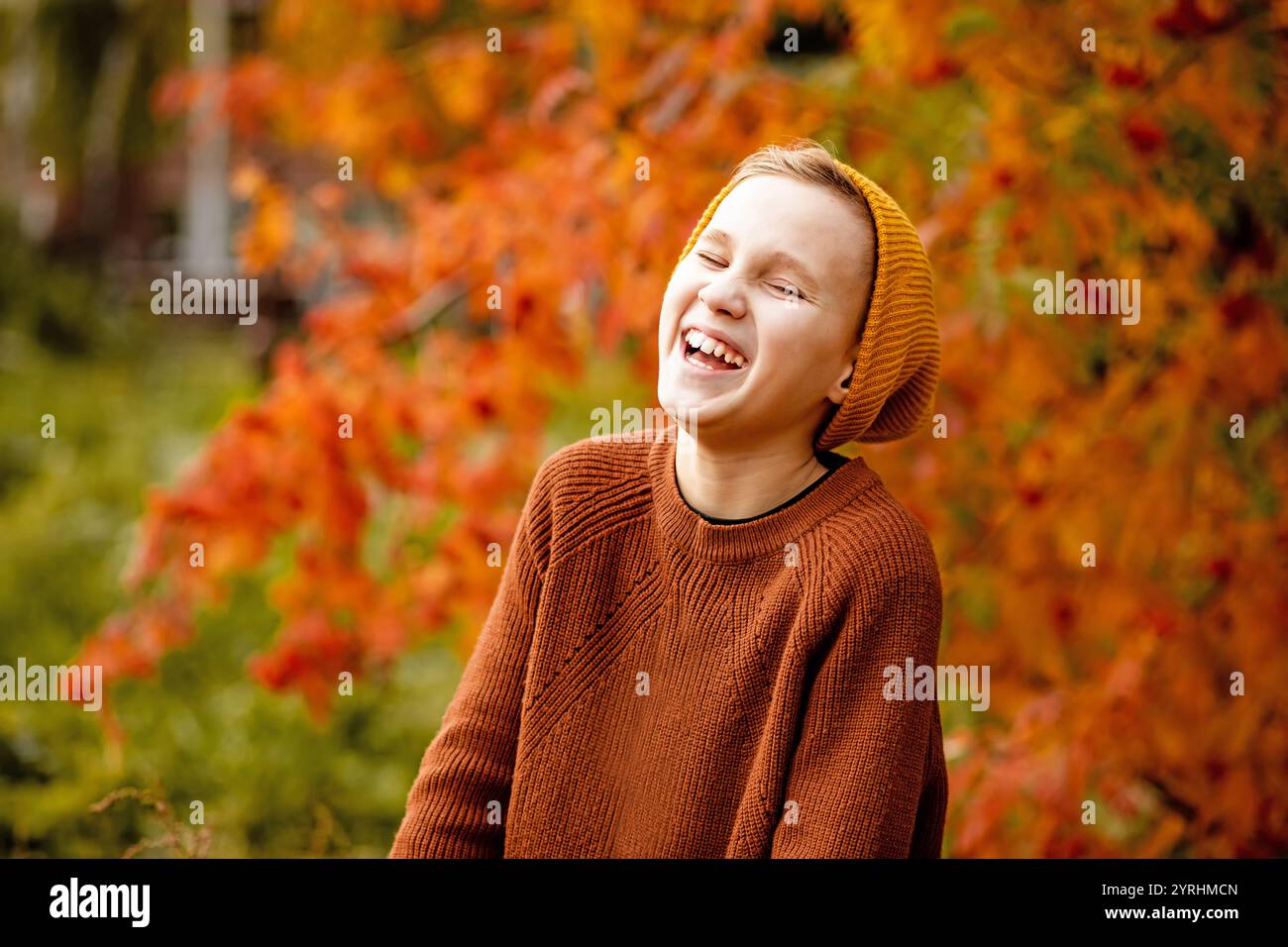 Un bambino allegro in un comodo maglione e berretto si diverte a ridere in mezzo alle vibranti foglie autunnali in un parco cittadino i colori caldi catturano l'essenza dell'autunno Foto Stock