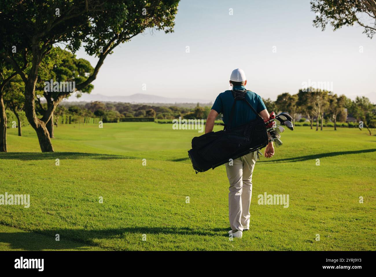 Uomo che cammina su un lussureggiante campo da golf portando mazze nella sua borsa da golf in un resort, godendosi il clima soleggiato e il paesaggio panoramico. Foto Stock
