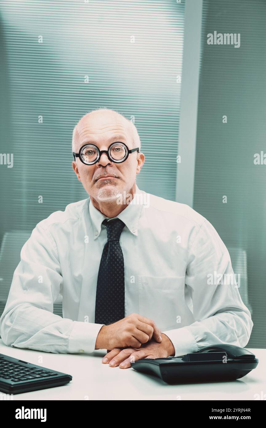 Uomo d'affari nerbo con occhiali spessi seduti alla sua scrivania, che guarda seriamente la fotocamera mentre è circondato dagli elementi essenziali dell'ufficio, trasmettendo una professione Foto Stock Uomo d'affari nerbo con occhiali spessi seduti alla sua scrivania, che guarda seriamente la fotocamera mentre è circondato dagli elementi essenziali dell'ufficio, trasmettendo una professione Foto Stock