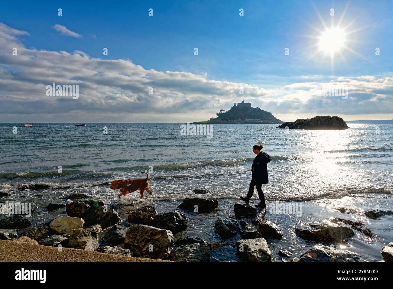 St. Michaels Mount in silhouette con il sole in un cielo blu con l'alta marea e una donna e un cane che camminano nel mare Marazion Cornovaglia Inghilterra Regno Unito Foto Stock