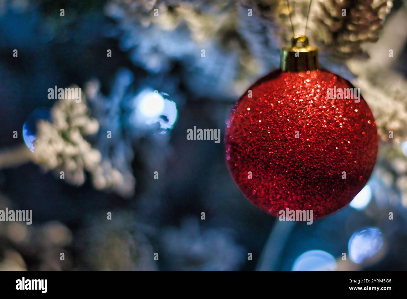 Uno scatto ravvicinato di un singolo ornamento natalizio rosso glitter appeso su un albero di Natale artificiale sfocato e innevato con blu e whi sfocati Foto Stock