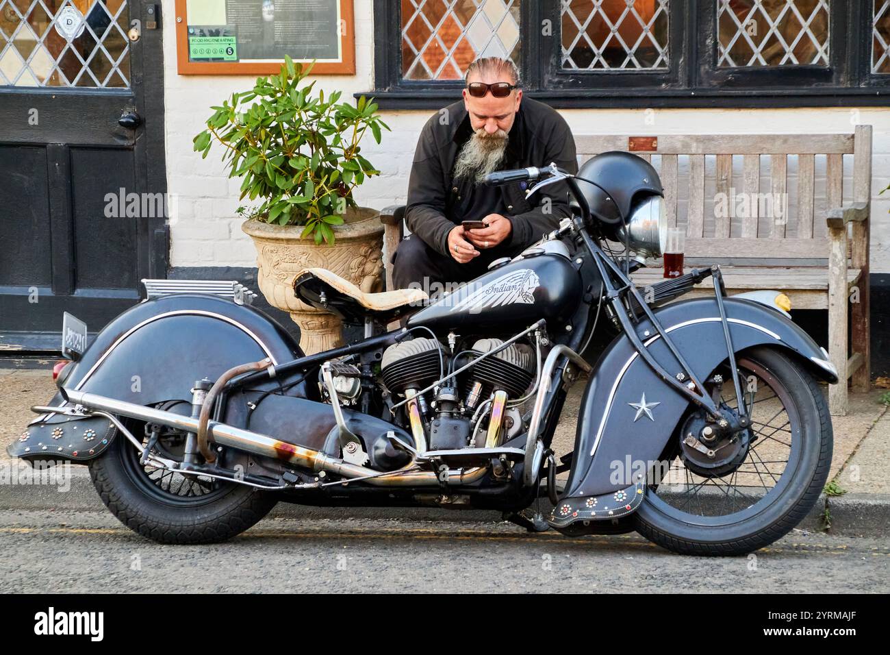 Una motocicletta nera Indian Chief del 1940 parcheggiata, con il pilota seduto su una panchina che beve un drink e guarda il suo cellulare. Foto Stock