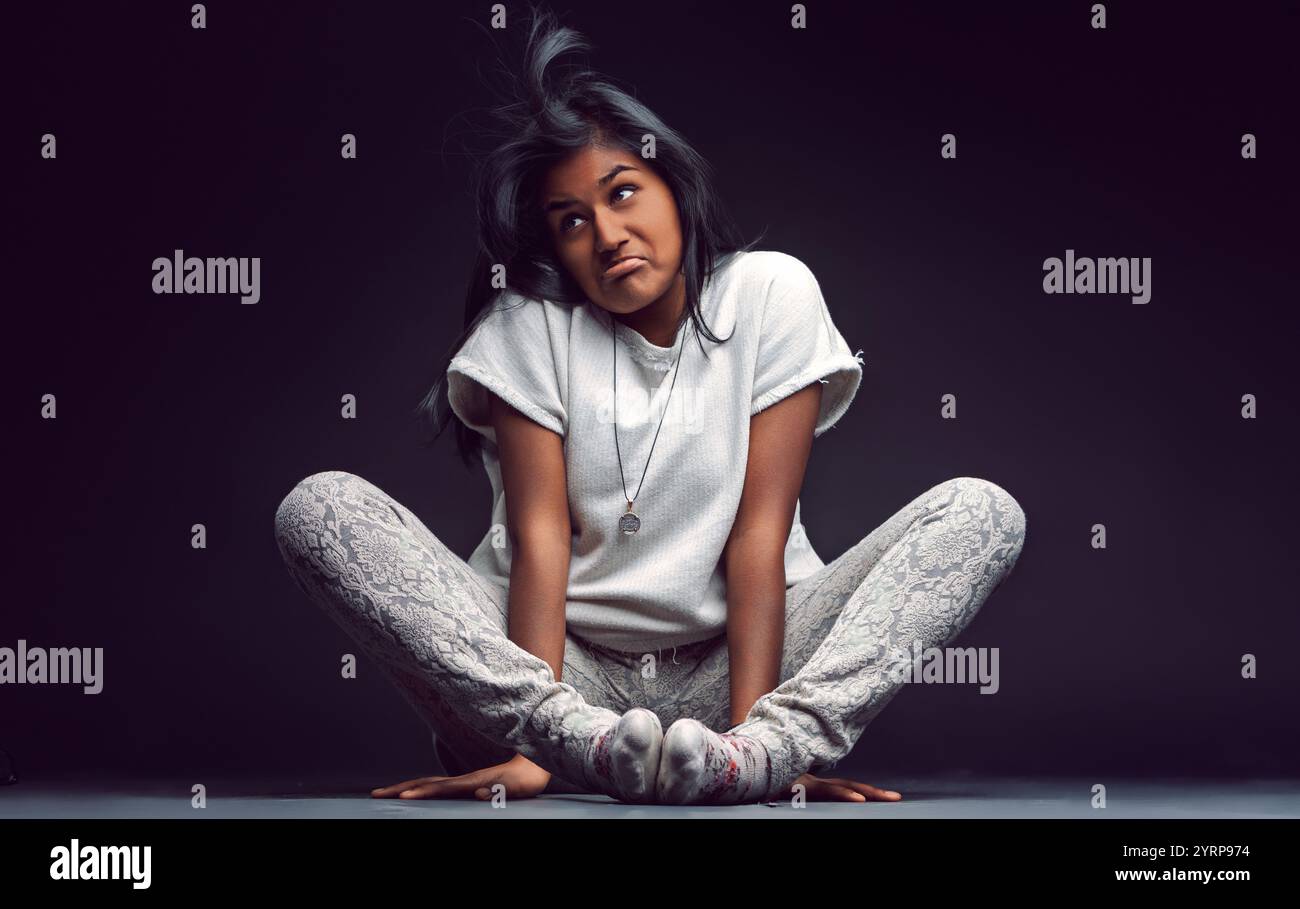 Giovane donna indiana seduta sul pavimento con le gambe incrociate, indossa calze bianche, pantaloni a fantasia e una t-shirt bianca, che crea un volto divertente mentre guardi Foto Stock