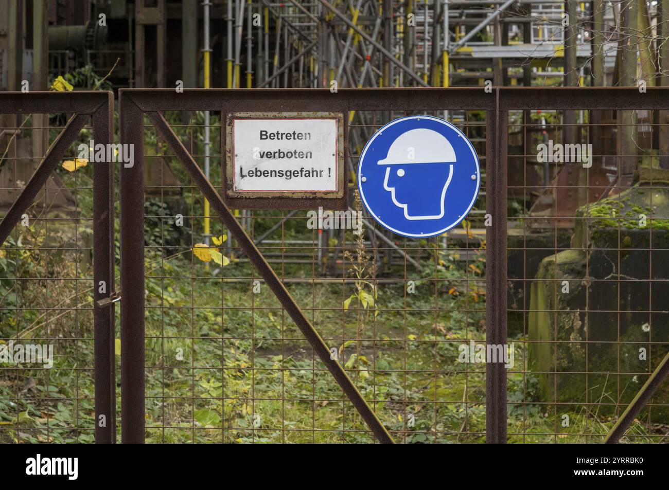 Cartelli di avvertenza in tedesco e cartelli obbligatori sul casco di fronte a un cancello arrugginito con un'area industriale con impalcature sullo sfondo Foto Stock