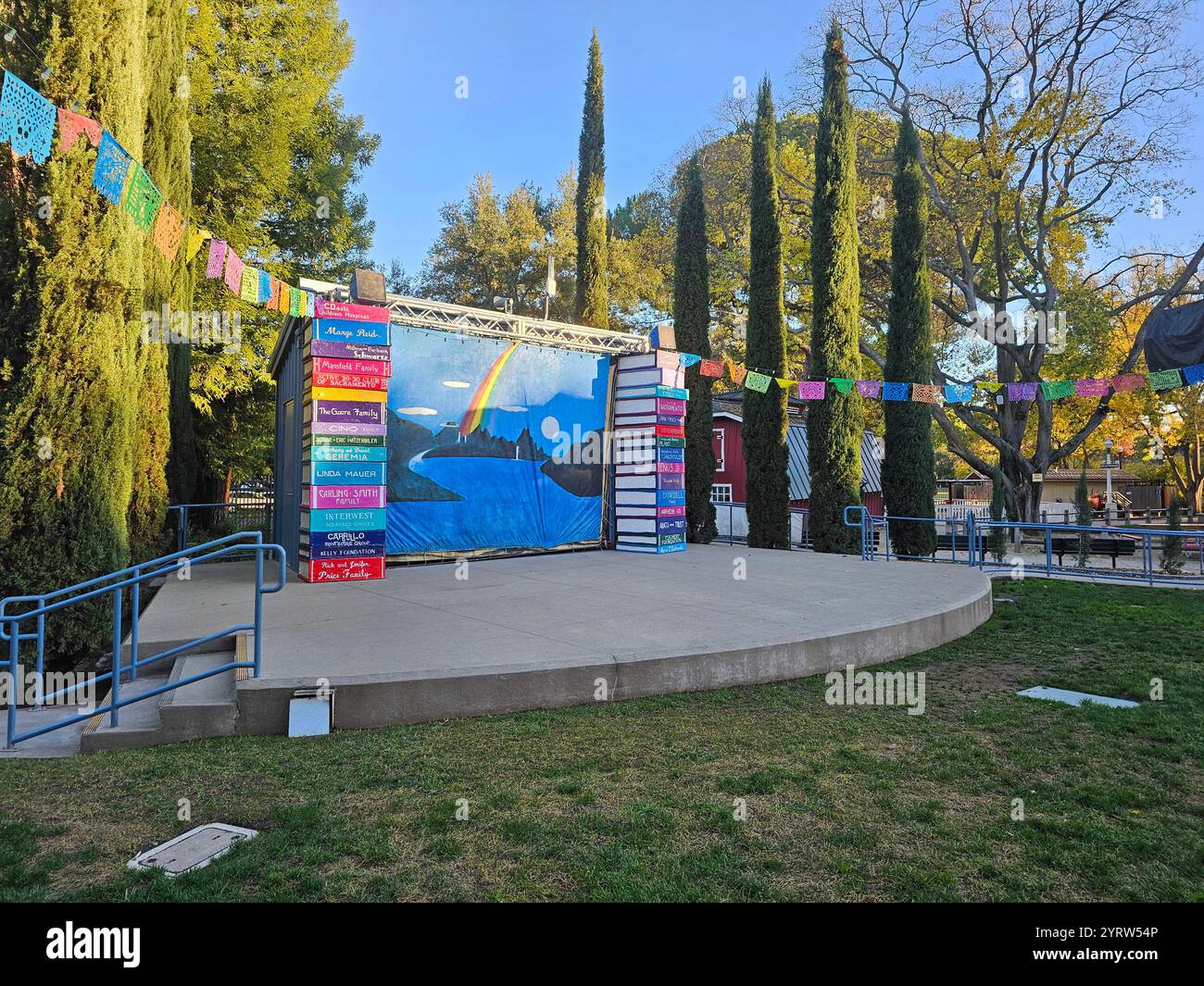 Palcoscenico all'aperto con decorazioni colorate, striscioni e sfondo arcobaleno dipinto circondato da alberi sotto un cielo azzurro. Foto Stock