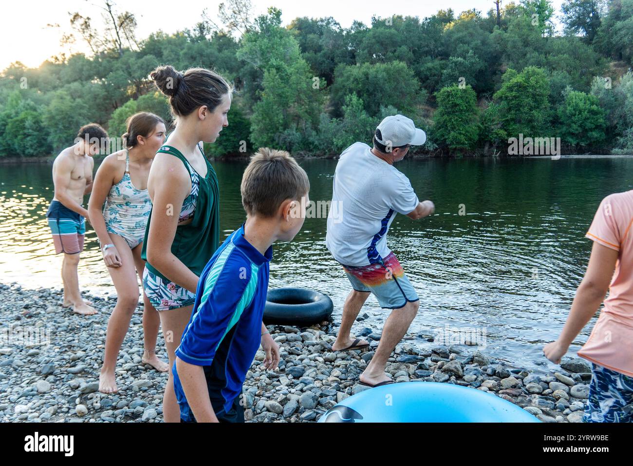 Il bambino lancia una pietra nel fiume mentre gli adulti osservano nelle vicinanze Foto Stock