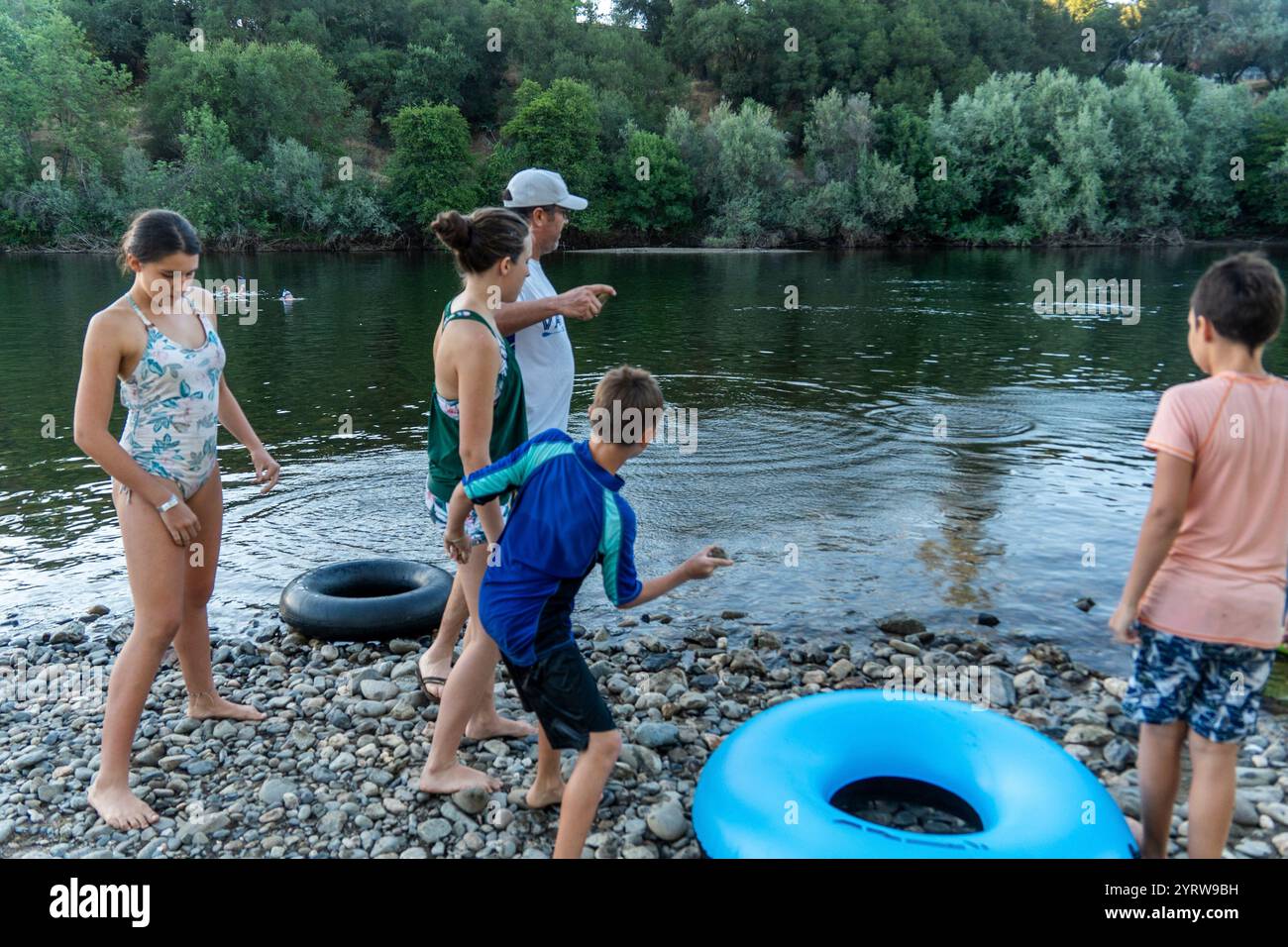 Il bambino lancia una pietra nel fiume mentre gli adulti osservano nelle vicinanze Foto Stock