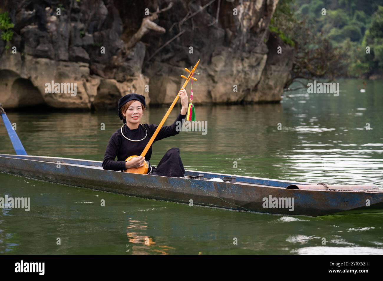 Smiling Tay Woman in una barca che suona lo strumento musicale Tinh sul lago Ba Be nel parco nazionale di Ba Be nella provincia di Bac Kan, Vietnam Foto Stock
