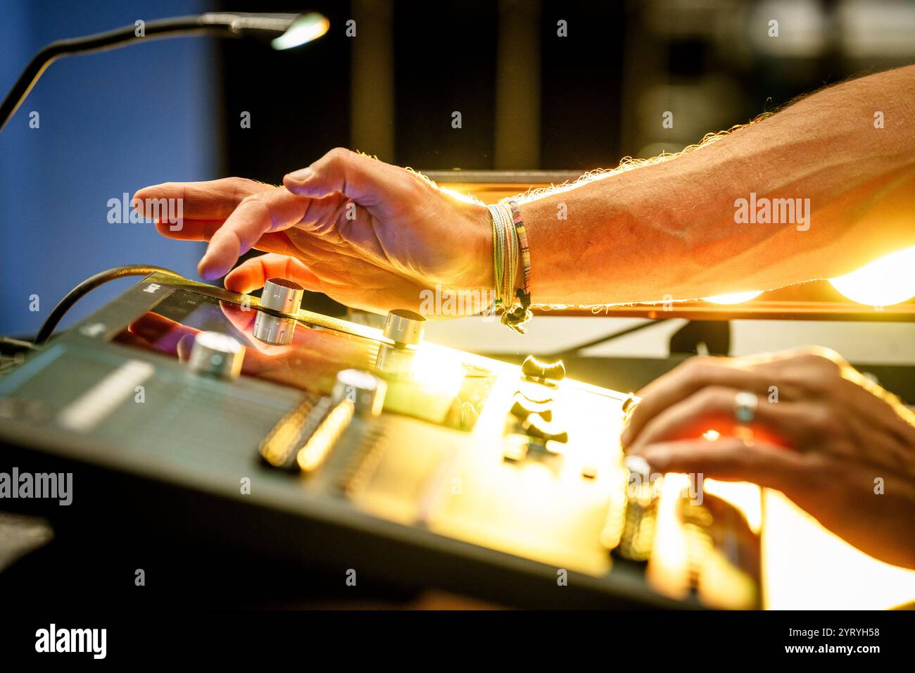 Primo piano delle mani di un tecnico che aziona una console di mixaggio audio, illuminata da luci di scena, che enfatizza la produzione audio in un evento dal vivo. Foto Stock