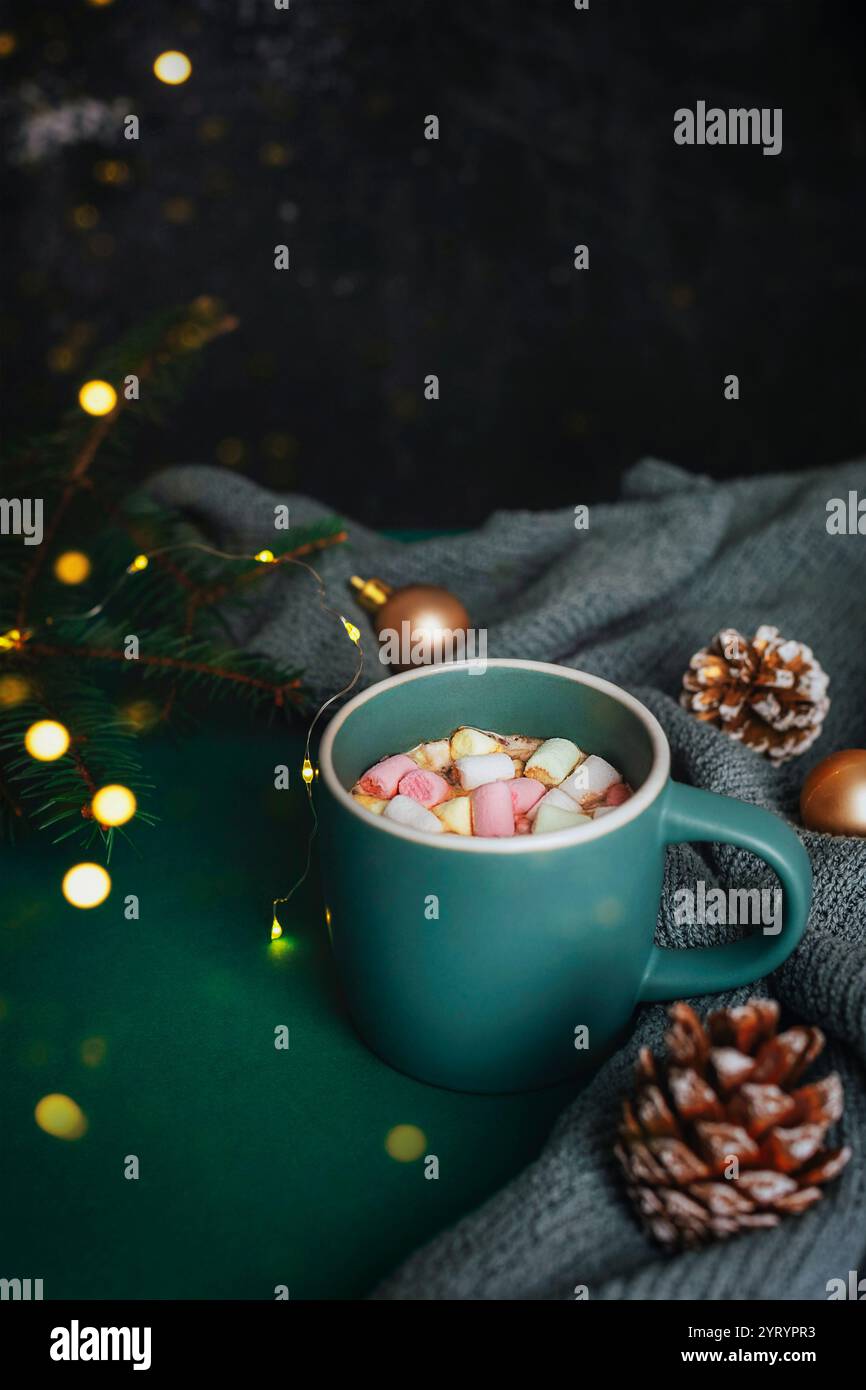 Tazza di cioccolata calda con marshmallow circondati da coni di pino, luci da favola e decorazioni natalizie su uno sfondo scuro e accogliente, creando un ambiente caldo e inviti Foto Stock