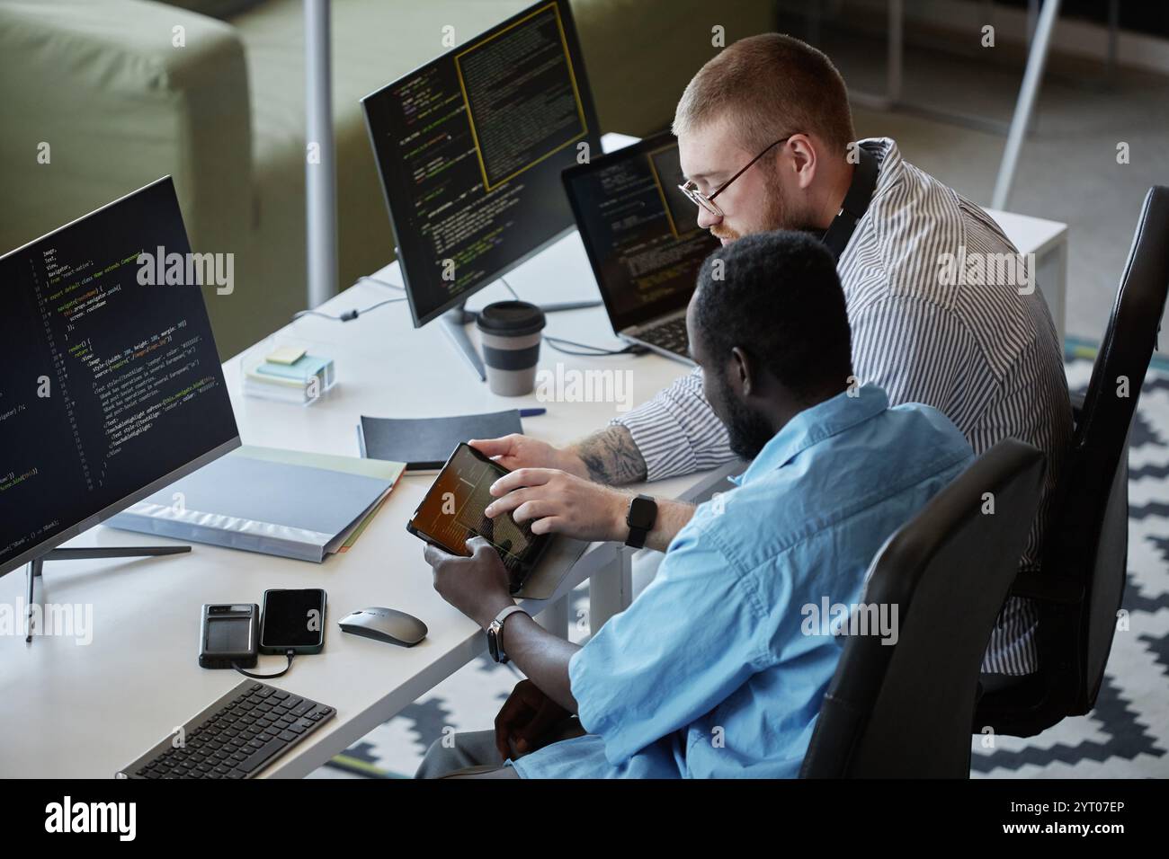 Due colleghi che lavorano insieme allo sviluppo di software in un moderno spazio di lavoro tecnologico, entrambi focalizzati sulla codifica e sull'utilizzo di strumenti digitali. Una persona sta mostrando informazioni su un tablet Foto Stock