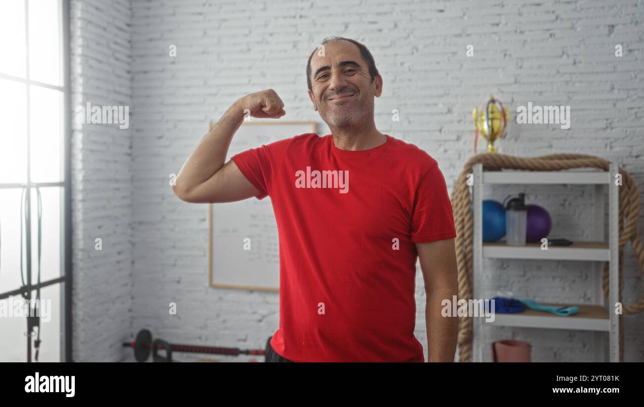 Uomo ispanico di mezza età che si flette braccio in palestra con pesi e trofeo sullo sfondo Foto Stock