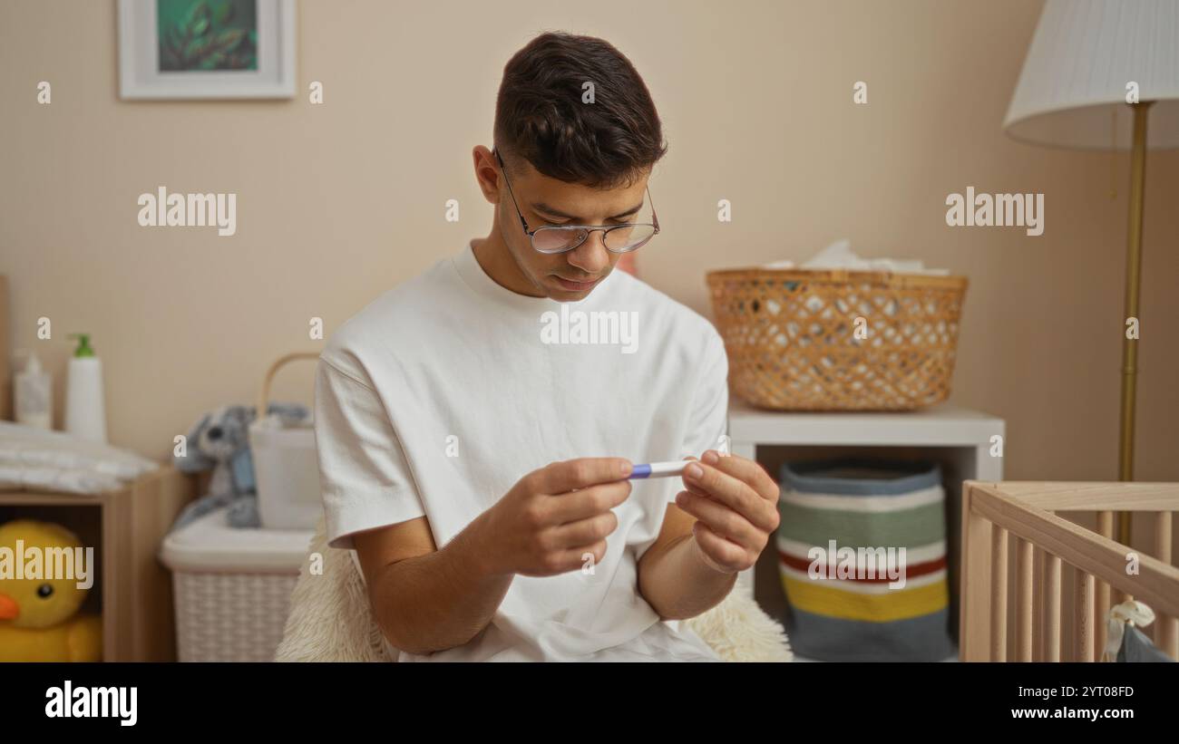 Il giovane nella stanza del bambino esamina il test di gravidanza circondato da un arredamento accogliente e culla, mostrando l'anticipazione e il calore nel momento genitoriale Foto Stock
