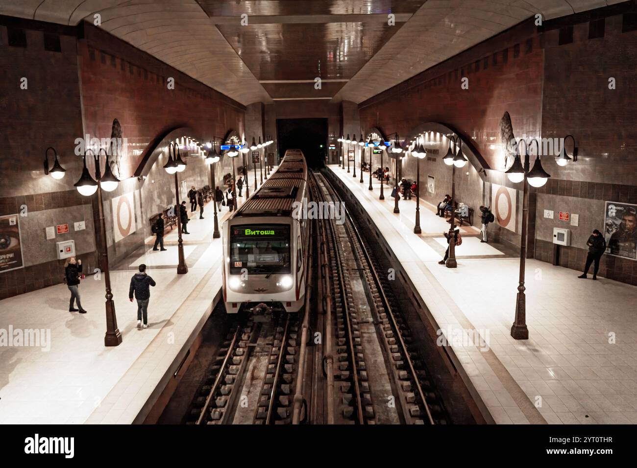Stazione della metropolitana di Lavov Most a Sofia, Bulgaria, un treno che arriva al binario, illuminato da lampade in stile vintage e circondato da pendolari. Foto Stock