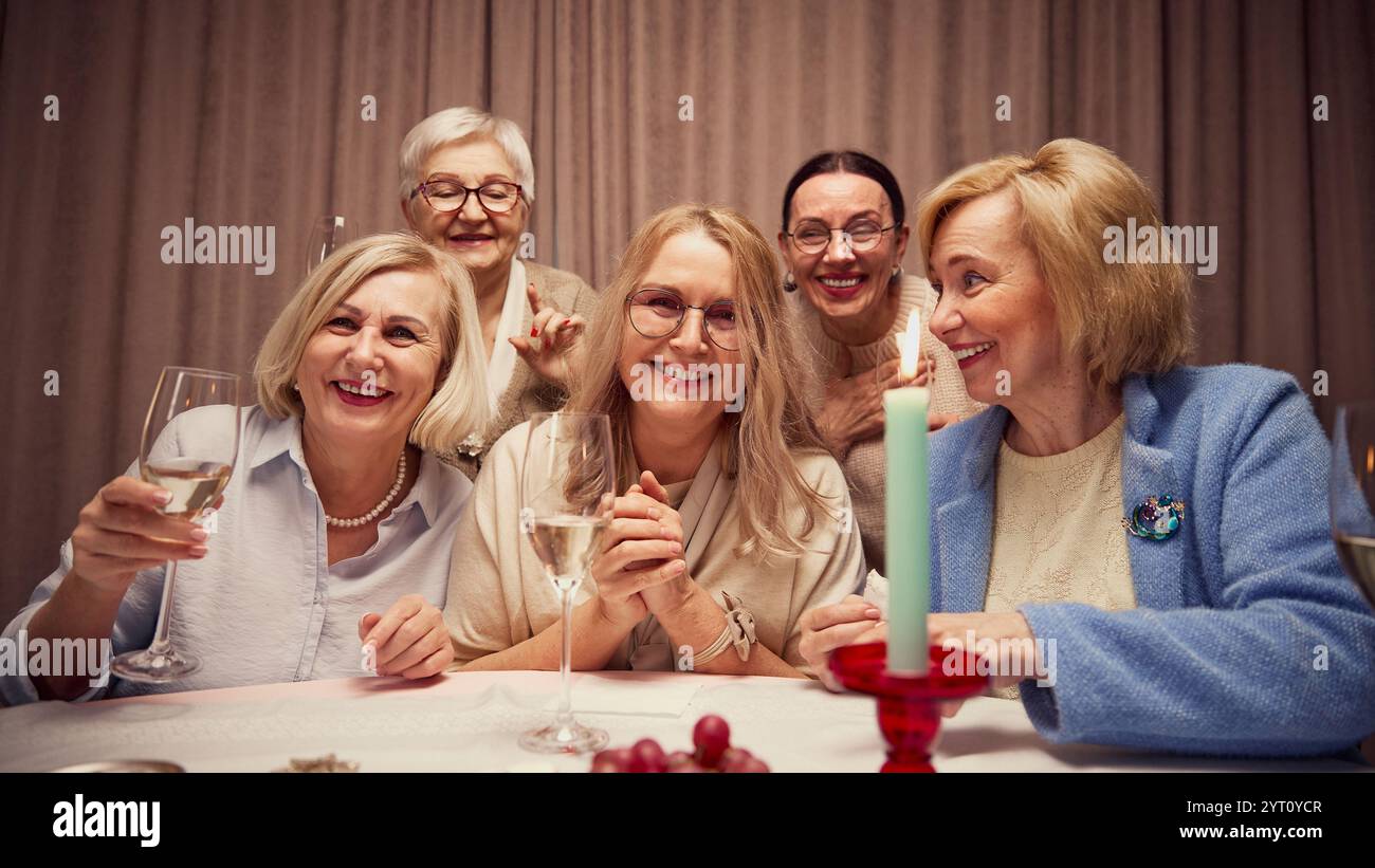 Gruppo di cinque anziane donne sorridenti, che si riuniscono intorno a un tavolo decorato con candele in serata accogliente, ridendo, parlando e bevendo vino Foto Stock