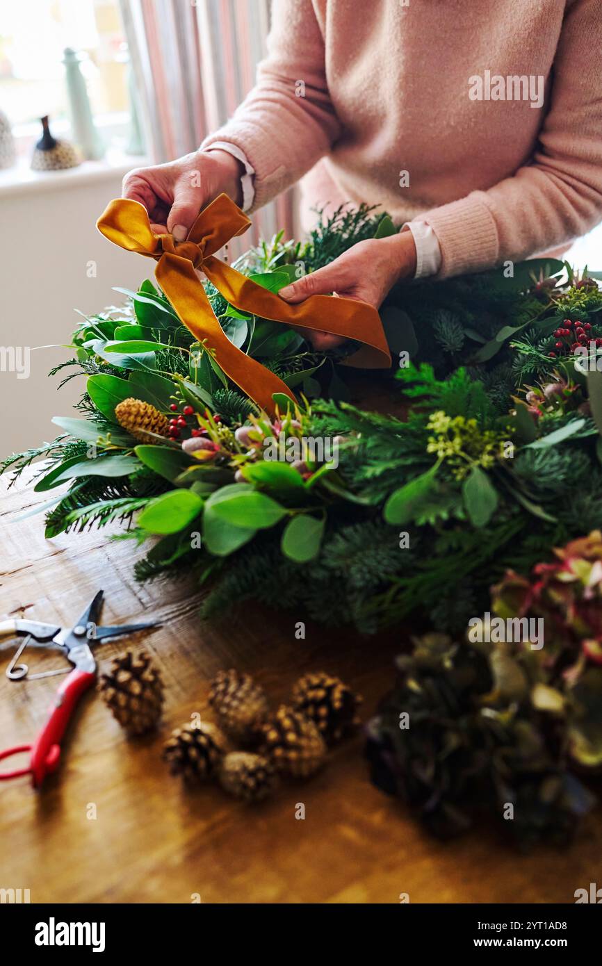 Primo piano di una donna che crea e decora le ghirlande natalizie su Table at Home Fastening Bow in nastro Foto Stock