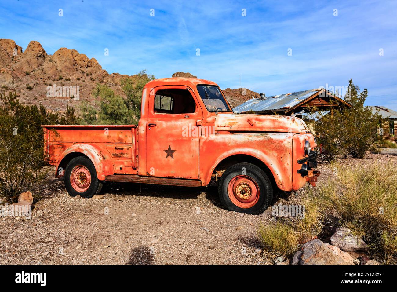 Un vecchio camion rosso arrugginito è parcheggiato in un deserto. Il carrello è ricoperto di ruggine e presenta una stella sul cofano. Il paesaggio desertico è arido e desolato, wi Foto Stock