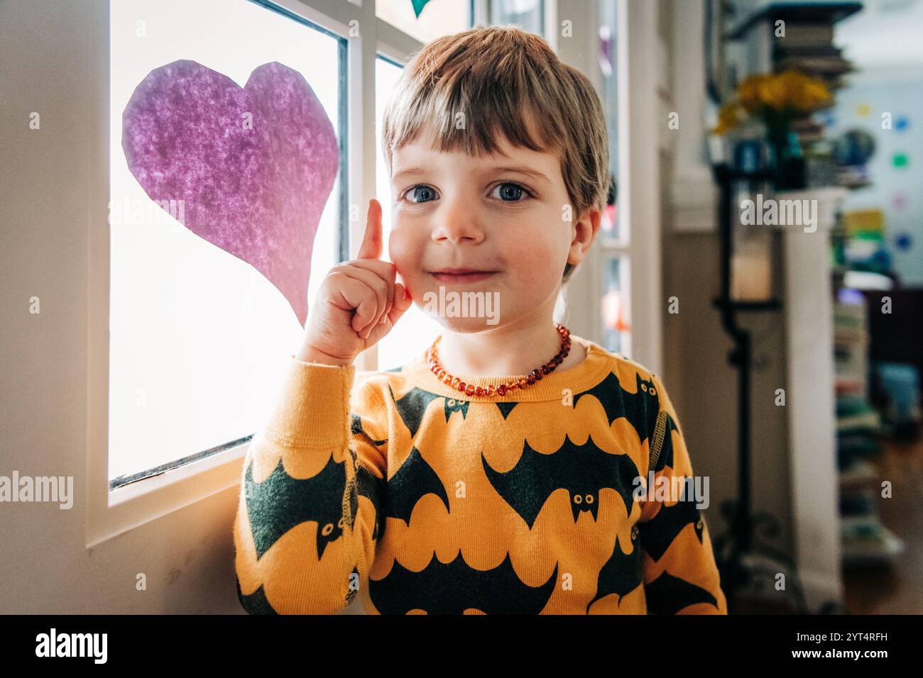 Bambino sorridente in pigiama con stampa a pipistrello vicino alla finestra decorata con il cuore Foto Stock