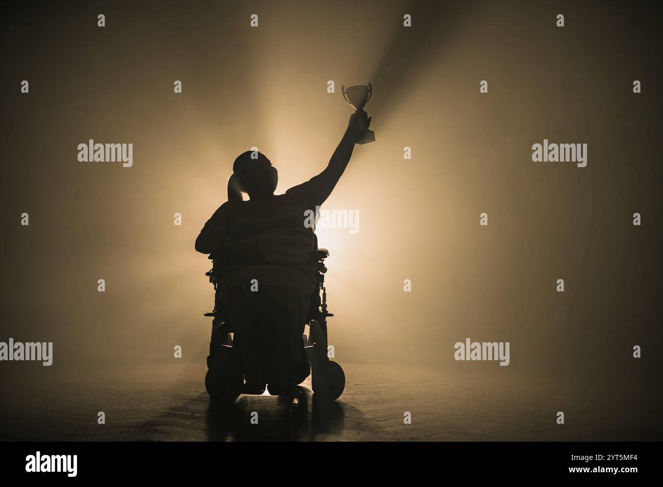 Donna con disabilità che riceve una coppa della vittoria mentre siede su una sedia a rotelle in una stanza buia con una retroilluminazione bianca. Foto Stock