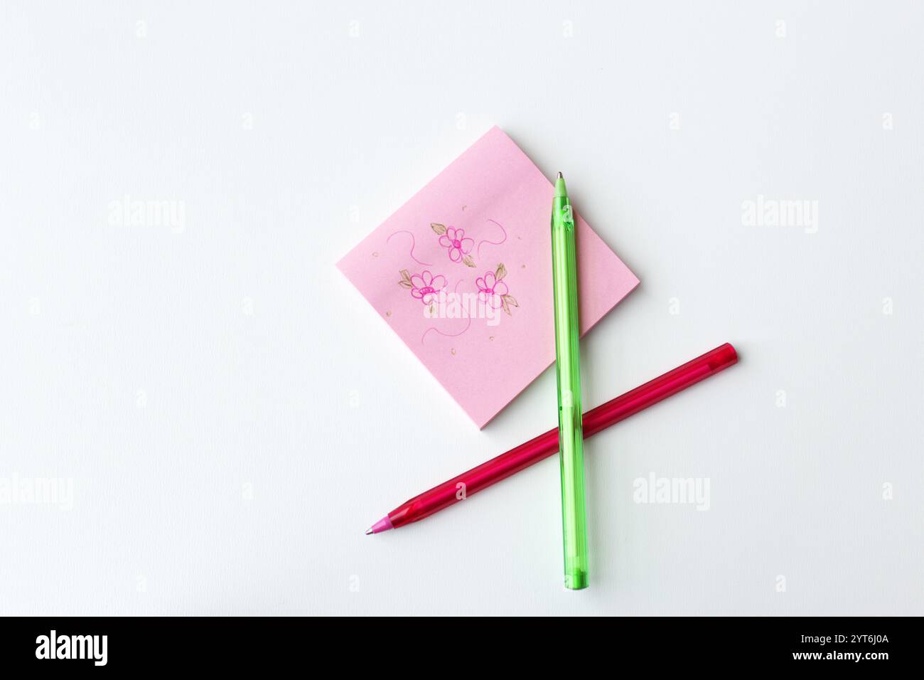 Penne a sfera di colore rosa e verde accanto a un blocco appiccicoso quadrato con fiori di doodle disegnati su di esso. Cancelleria creativa per lezione d'arte. Foto Stock
