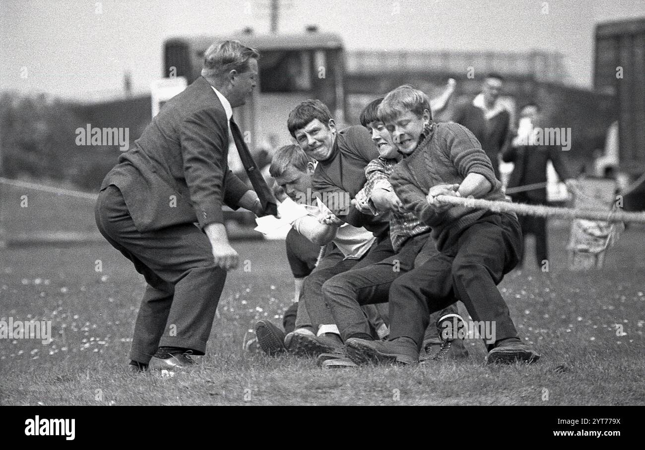 1968, storico, all'esterno in un campo di una fayre o di una fete della contea, una squadra di ragazzi che tira forte su una corda in una gara di tiro alla fune, un allenatore maschio con vestito e cravatta accanto a loro che dà istruzioni e supporto, Inghilterra, Regno Unito. Foto Stock