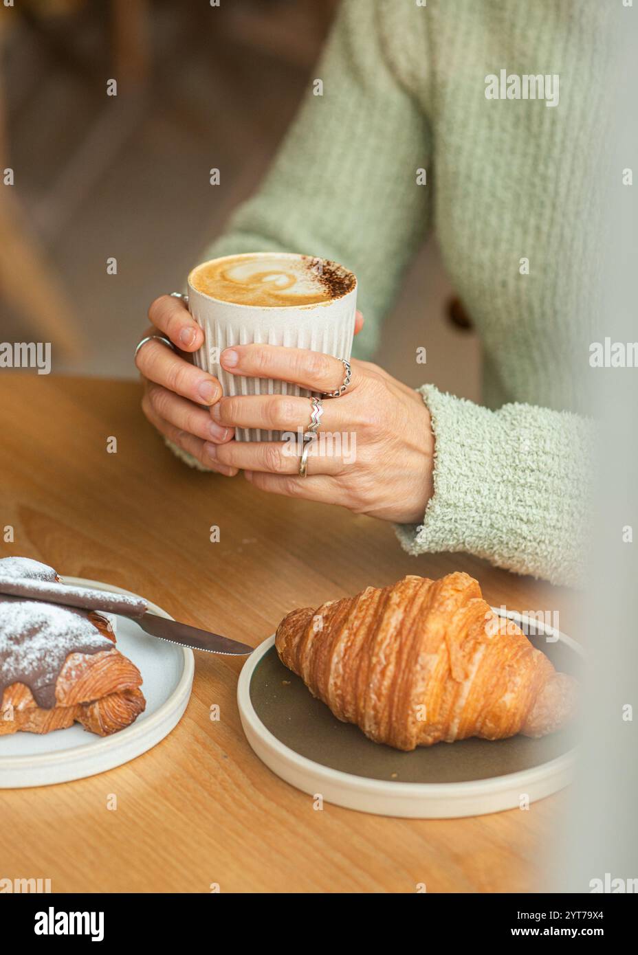 Un'accogliente caffetteria con una persona in un maglione verde che tiene un cappuccino in una tazza costolata. Sul tavolo di legno, un croissant al cioccolato. Foto Stock