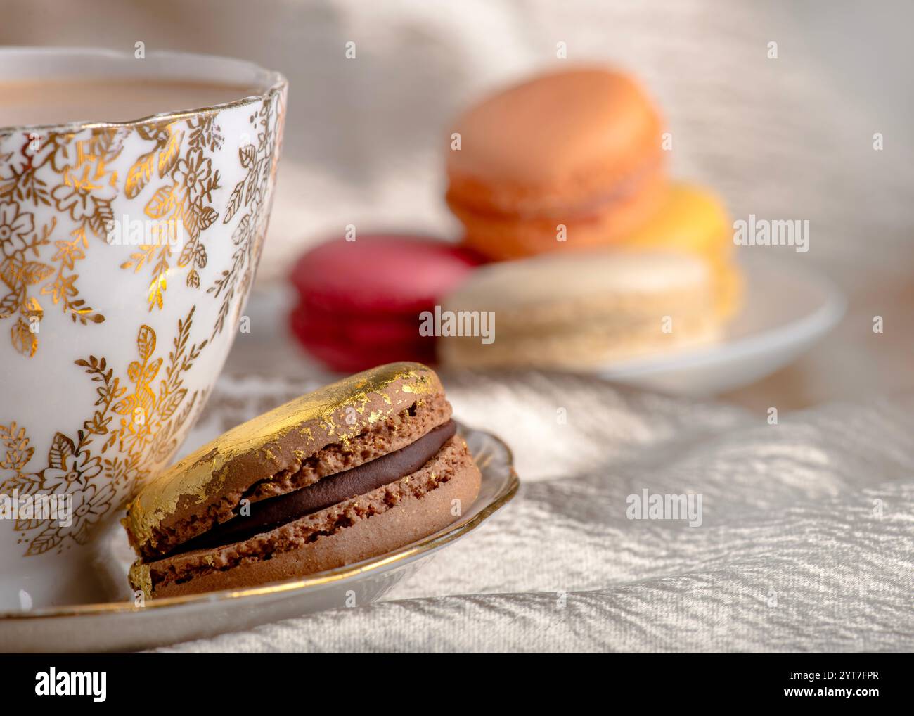 scena lussuosa con un tè rifinito in oro abbinato a un macaron al cioccolato impreziosito da foglie d'oro, circondato da macaron colorati su un testo morbido Foto Stock