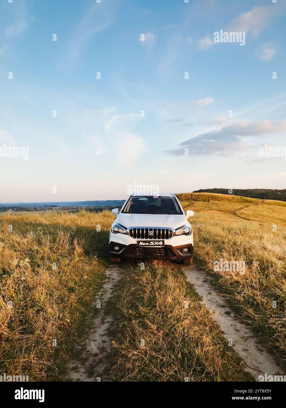 White Suzuki SX-4 sul sentiero in cima alla catena montuosa. Vista frontale del SUV giapponese in pista nei prati collinari della campagna al tramonto. Foto Stock