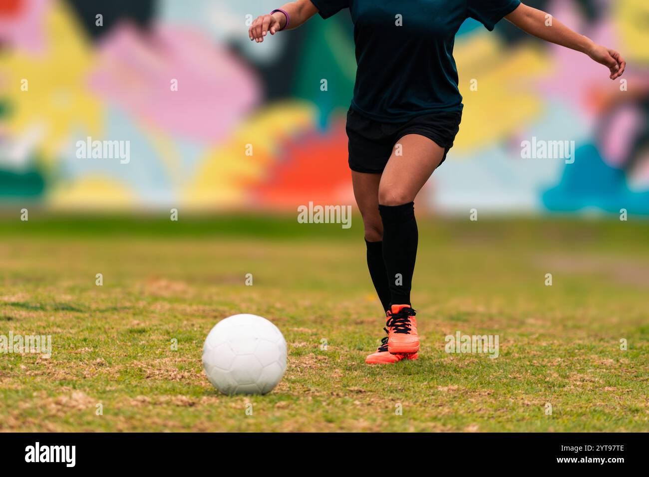 Vista delle gambe della bruna giocatrice di calcio che corre per calciare la palla. su un'erba verde all'aperto, con sfondo colorato. con abbigliamento sportivo. Foto Stock