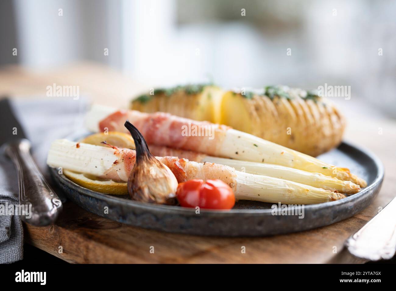 Asparagi bianchi fritti avvolti in pancetta con patate alle erbe e pomodori su piatto di ceramica nera e tavolo di legno. Profondità di campo ridotta. Foto Stock